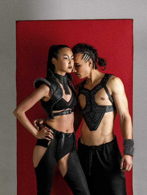Man and woman against a red background wearing leather beaded body pieces and black pants.