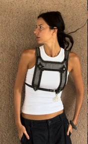 Woman wearing a black mesh harness over a white tank top against a textured beige wall.
