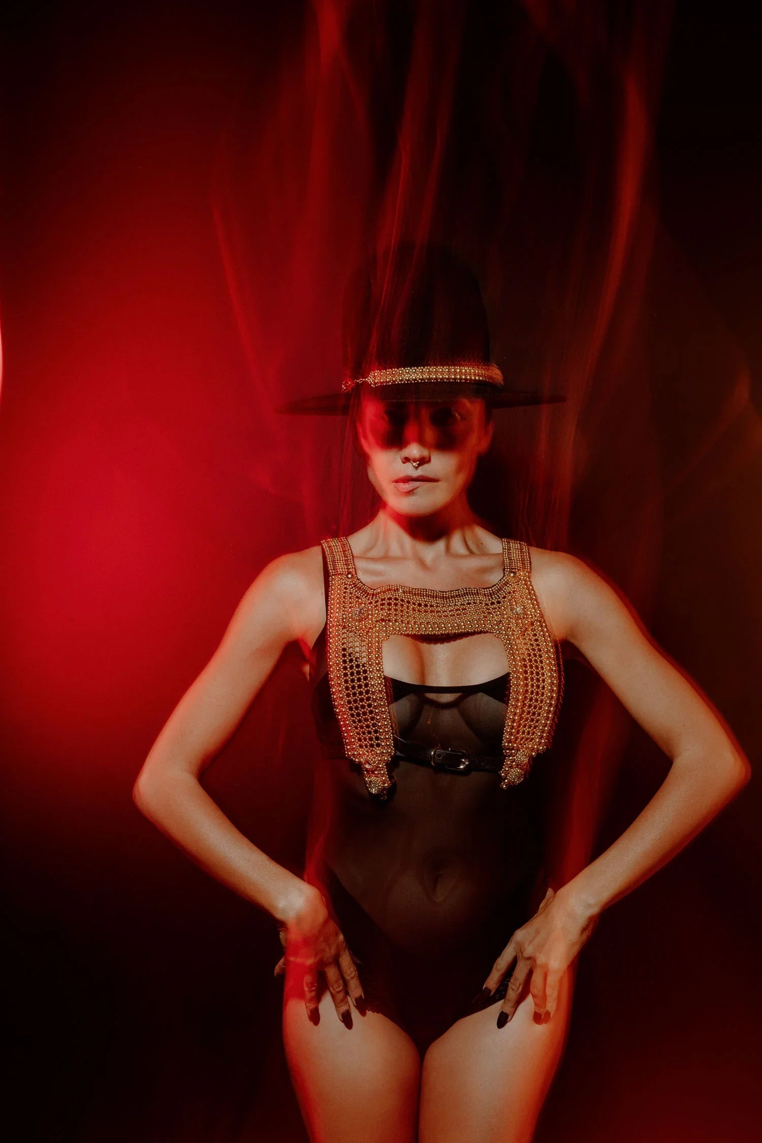 Woman wearing a black hat with gold beaded hatband and one piece swimsuit with a gold beaded harness over top against a red background