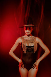 Woman wearing a black hat with gold beaded hatband and one piece swimsuit with a gold beaded harness over top against a red background