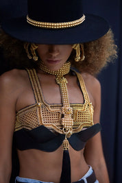Woman wearing a gold beaded necklace and bra top and black hat with a dark background