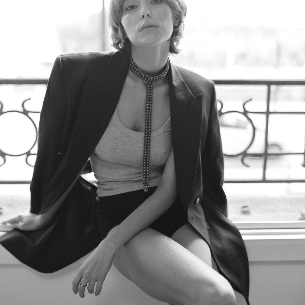 Woman wearing a black blazer and slim t-choker necklace by object & Dawn sitting on a ledge.