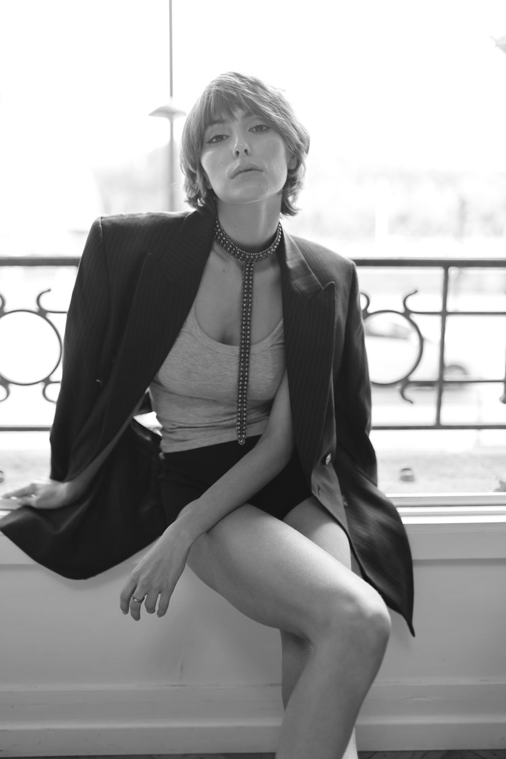 Woman wearing a black blazer and slim t-choker necklace by object & Dawn sitting on a ledge.