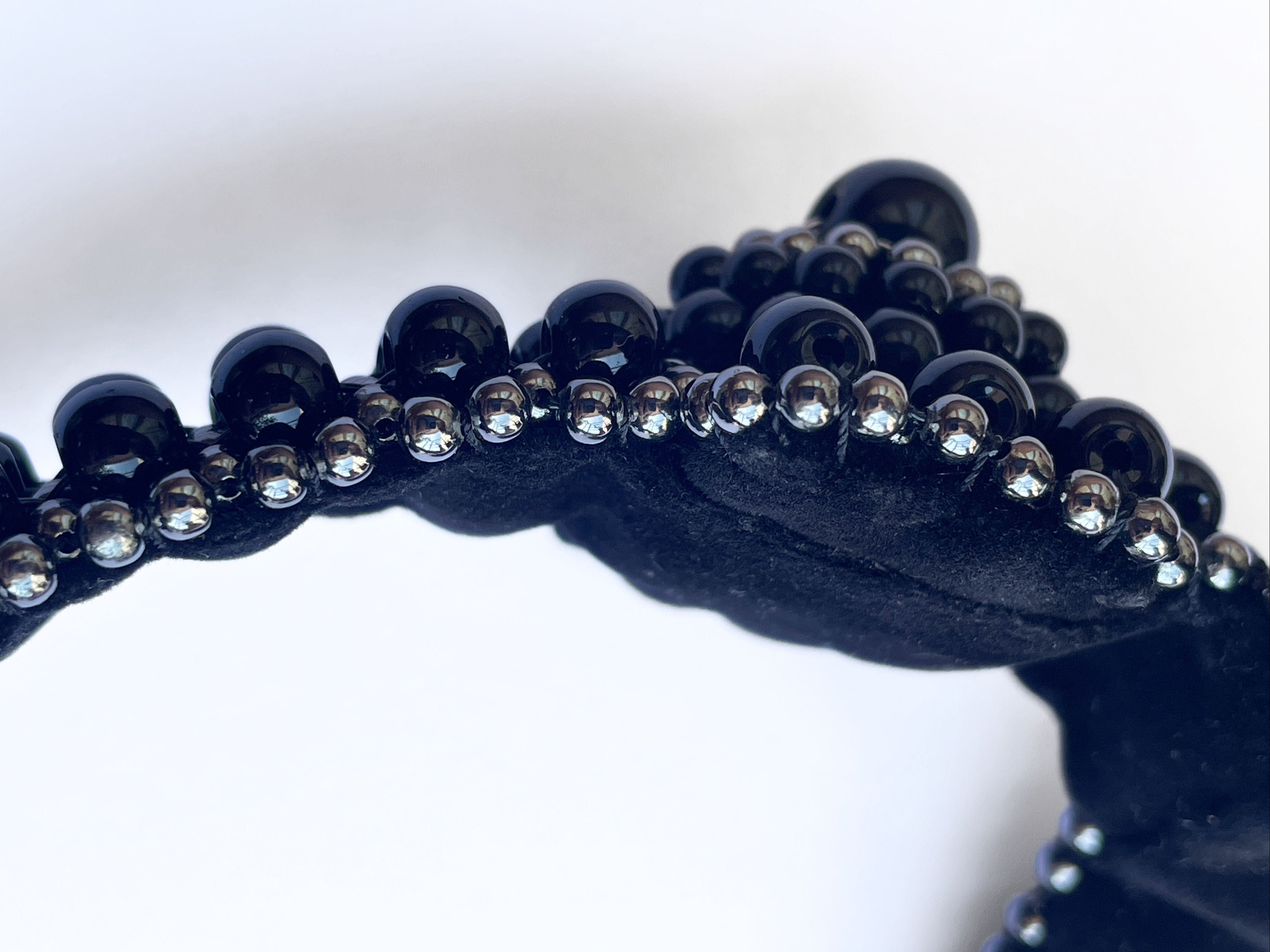 A close-up of a black beaded accessory, likely a headband or trim, featuring large glossy black beads and smaller clear rhinestone-like beads on a white background.