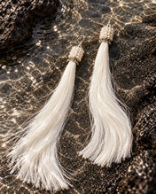 A pair of white 22 inch tassels with pearl beaded caps lying on wet black sand and lava rock