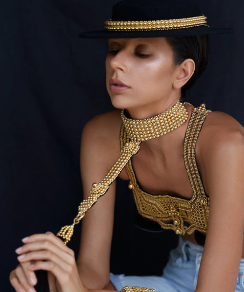 Woman wearing gold beaded jewelry by Object & Dawn and a black hat against a dark background